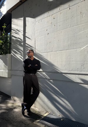Hombre vestido de negro y con gafas de sol apoyado contra una pared de hormigón gris iluminada por el sol, con patrones de sombra.