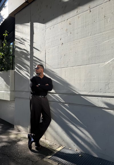 Hombre vestido de negro y con gafas de sol apoyado contra una pared de hormigón gris iluminada por el sol, con patrones de sombra.