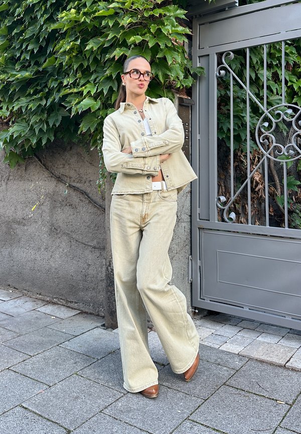 Joven mujer con gafas, vestida con chaqueta vaquera beige y pantalones, de pie con los brazos cruzados junto a una puerta metálica gris y una pared cubierta de hojas.