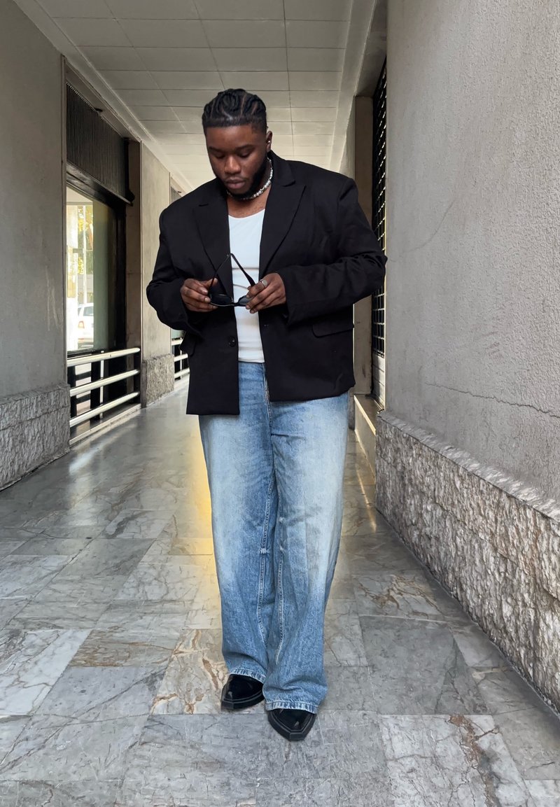 Black blazer worn over a white shirt, complemented by light blue distressed jeans and black shoes. Holding sunglasses while standing on marble flooring.