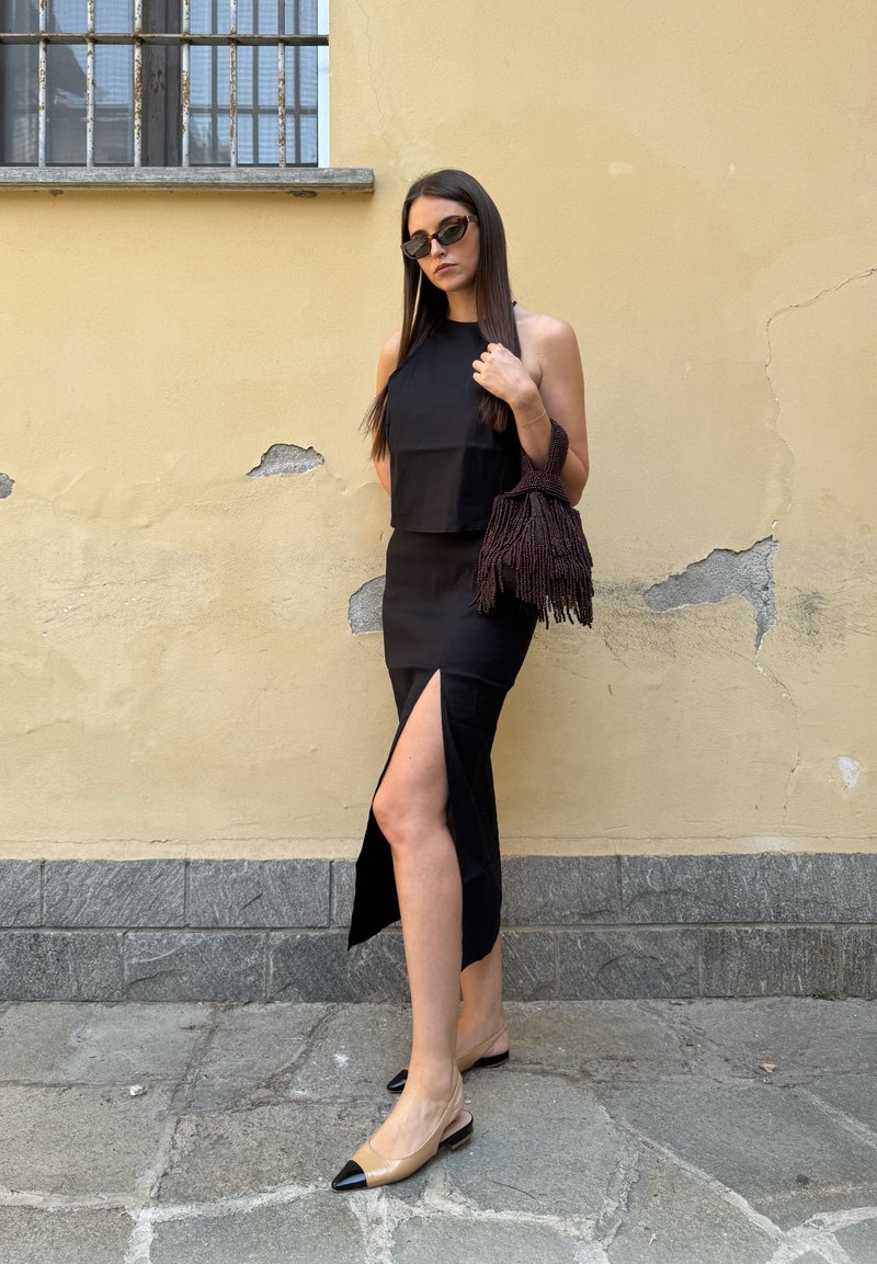 Femme en haut noir sans manches et jupe fendue tient un sac foncé à franges, porte des lunettes de soleil et des ballerines beiges à bouts noirs, debout près d'un mur jaune.