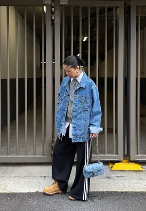 Persona que lleva una chaqueta vaquera sobredimensionada, suéter gris, camisa blanca, pantalones negros con rayas blancas, botas marrones y sostiene un bolso azul acolchado.