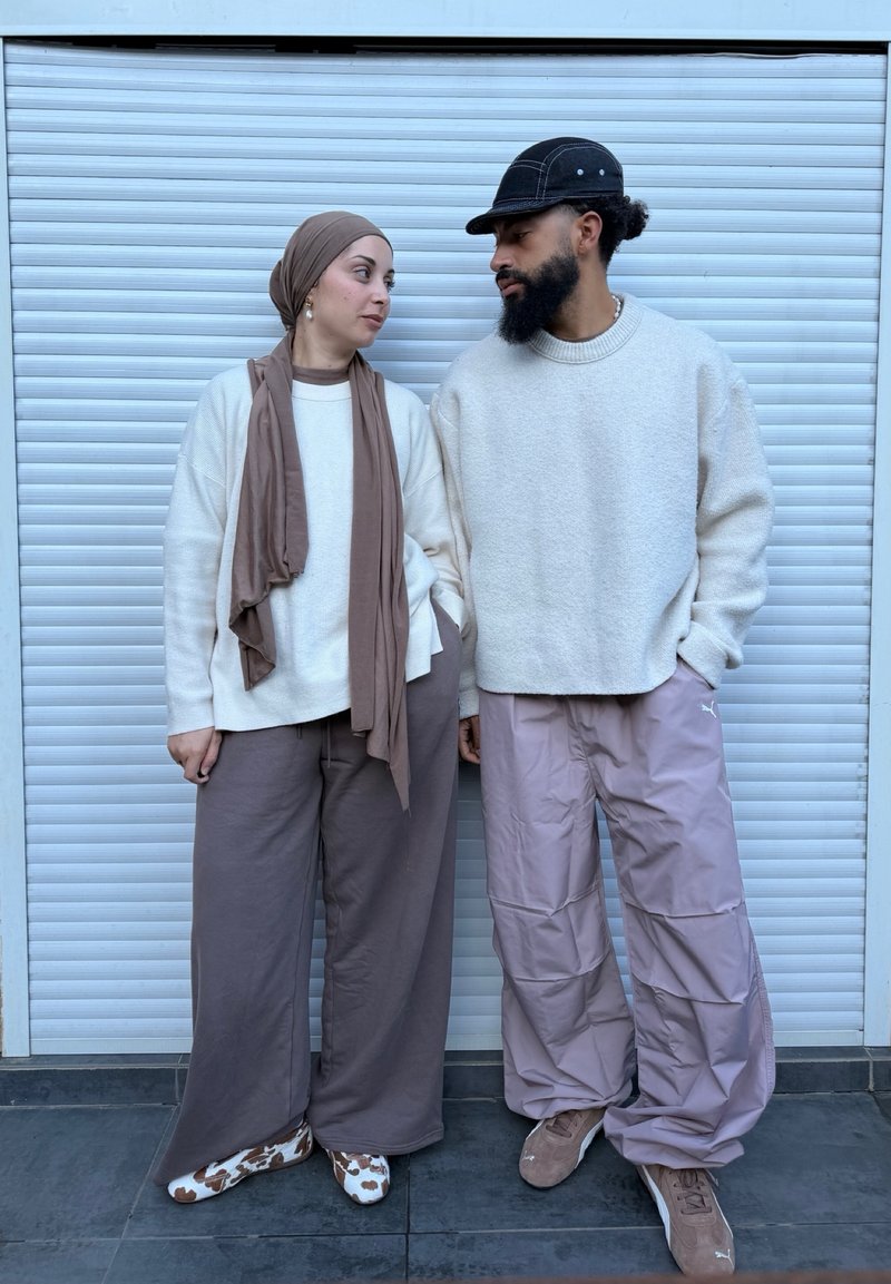 Deux personnes se tiennent côte à côte devant un volet blanc fermé ; l'une porte un foulard beige et un pantalon, l'autre une casquette noire et un pantalon violet clair.