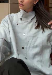 Veste oversize bleu clair avec col montant, boutons-pression, poche latérale et texture lisse ; portée sur un pantalon foncé.