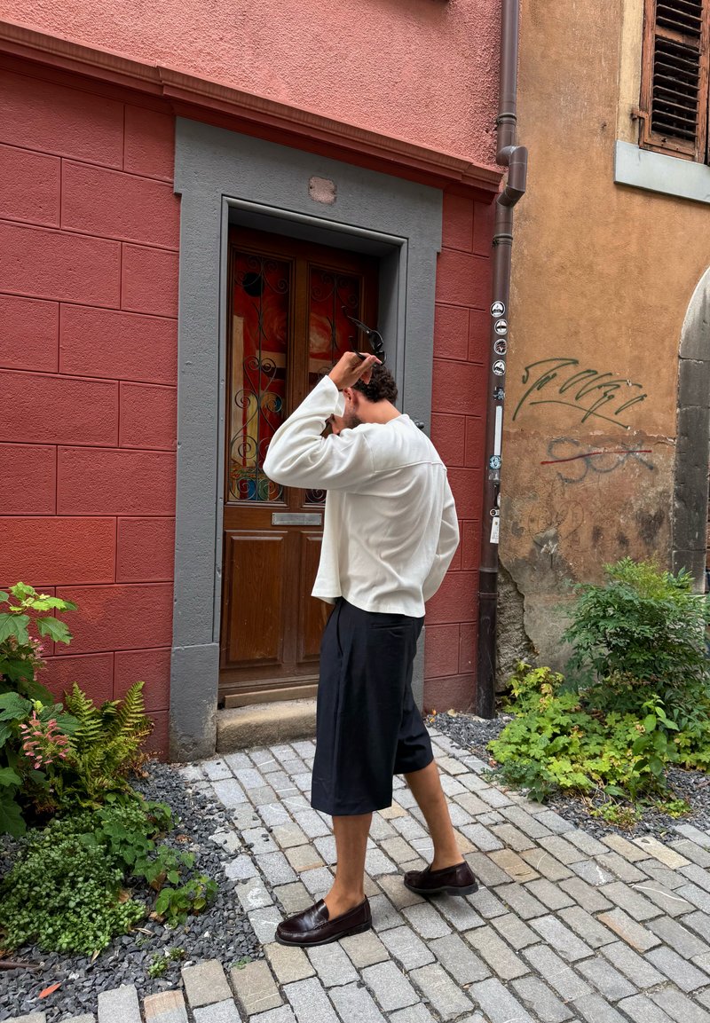 Personne en chemise blanche à manches longues et short noir ajustant des lunettes de soleil près d'une porte en bois sur un chemin de pierre avec des plantes à proximité.