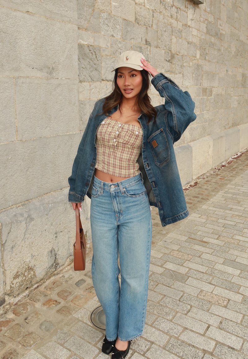 Jaqueta de ganga sobre um top de xadrez cropped, jeans de pernas largas azul claro, sapatos planos pretos e um boné bege com logotipo. Bolsa castanha na mão.
