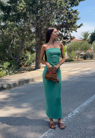 Vestido maxi de cuerpo ajustado en color verde azulado, con un diseño sin tirantes, tejido texturizado y fruncido. Accesorios incluyen un pequeño bolso marrón y sandalias planas.