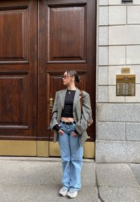 Blazer oversize a scacchi, top corto nero, jeans azzurri chiari, sneakers grigie; in piedi di fronte a porte doppie in legno con dettagli in ottone.