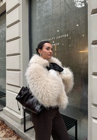 Fluffy beige faux bontjas met brede mouwen, zwarte leren handschoenen en donkergroene geribde broek. Zwarte tas met aangespannen trekkoorden.