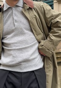 Bărbat îmbrăcat într-un trench coat bej peste un pulover gri cu nasturi și pantaloni negri, stând în aer liber lângă trepte din piatră și fațada unei clădiri.