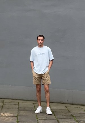 Camiseta corta de manga azul claro con el texto "LE JARDIN", pantalones cortos beige y zapatillas blancas, de pie sobre pavimento de piedra contra una pared gris.