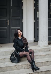 Pull noir, chemise blanche à col, jupe plissée noire, collants noirs à motif, bottes plateformes noires, et un sac à main rond noir sur des marches en pierre.