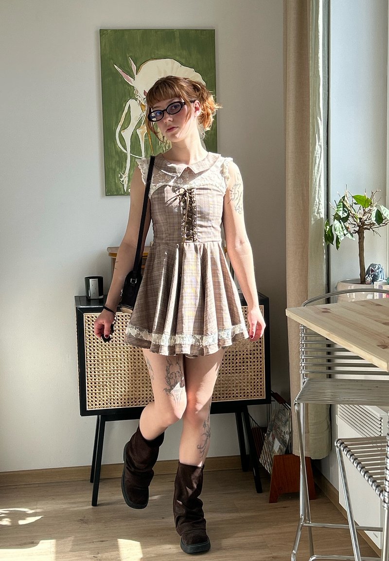 Jeune femme avec des lunettes portant une robe beige à lacets et des bottes foncées, debout dans une pièce ensoleillée avec des meubles et une plante.
