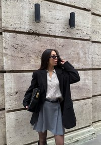 Blazer noir surdimensionné, pull en maille torsadée gris clair, jupe grise et lunettes de soleil noires. Porte une pochette noire avec des accents dorés.