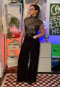 Femme portant un haut à imprimé léopard et un pantalon noir taille haute, debout à l'intérieur près d'une étagère avec des plantes, des chaussures et des posters colorés.