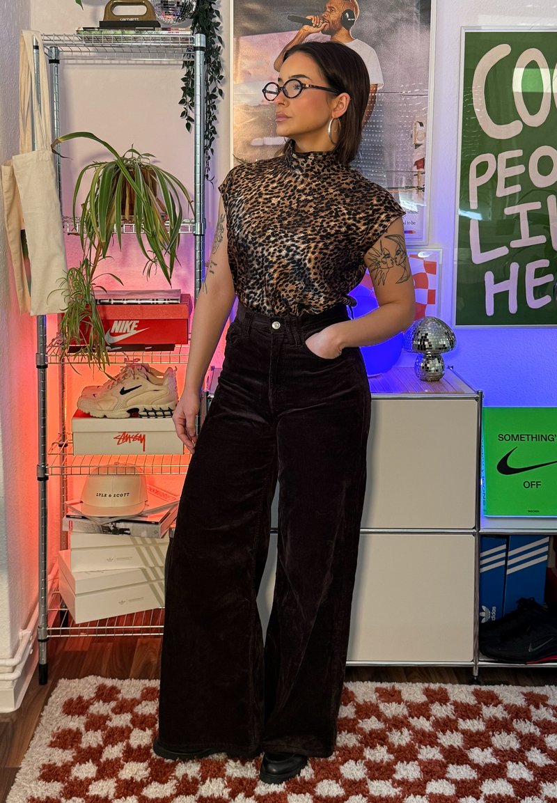 Femme portant un haut à imprimé léopard et un pantalon noir taille haute, debout à l'intérieur près d'une étagère avec des plantes, des chaussures et des affiches colorées.