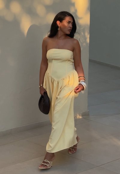 Vestido amarillo sin mangas con un corsé ajustado y falda fruncida. Acompañado de un bolso oscuro y sandalias beige con detalles atados.