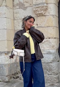 Brown leather bomber jacket, pale yellow knitted sweater, blue high-waisted jeans, and a small white crossbody bag with a playful detail.