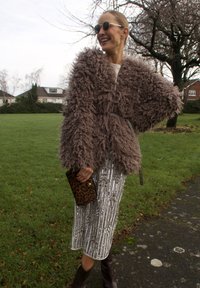 Manteau en fausse fourrure marron avec une texture duveteuse, associé à une jupe en sequins argentés et une pochette imprimé léopard. Des lunettes de soleil complètent la tenue.