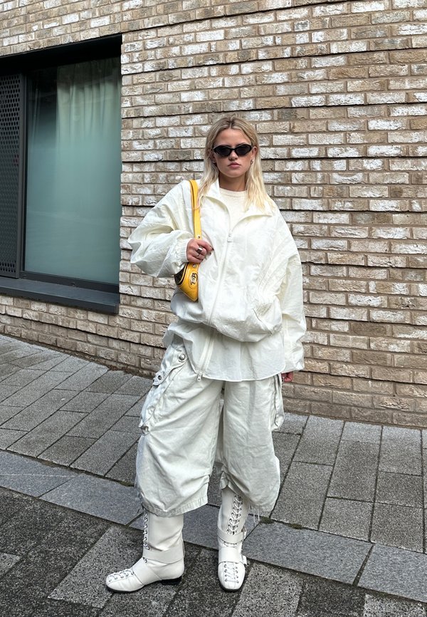 Jeune femme portant une veste blanche surdimensionnée, un pantalon cargo ample, des bottes blanches à lacets, des lunettes de soleil noires et un sac à épaule jaune, debout près d’un mur en brique.