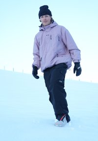 Pink waterproof jacket, black snow trousers, and gloves, worn while walking on snow; features a logo and a loose fit for ease of movement.