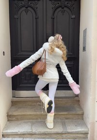 Fehér műszőrme-béléses dzseki, rózsaszín bolyhos kesztyűk, krémszínű bokacsizma, fehér leggings, és barna táska, sötét ajtó mellett lépcsőn pózolva.