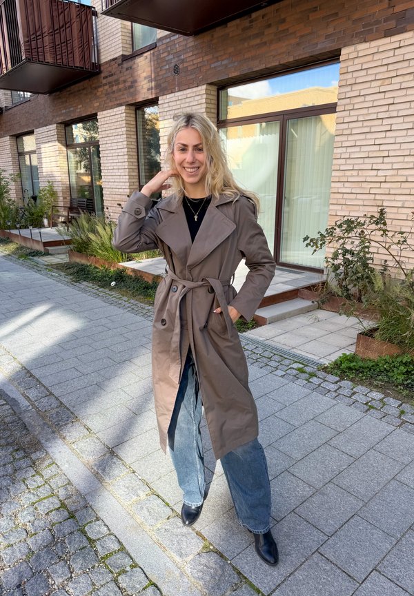 Joven mujer con cabello rubio que lleva un abrigo marrón tipo trench, top negro, vaqueros y botas negras, de pie y sonriendo en una acera urbana pavimentada.