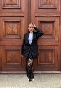 Blazer oversize negro con solapas, superpuesto sobre una blusa azul clara, combinado con pantalones de encaje negros con patrones florales y tacones blancos.
