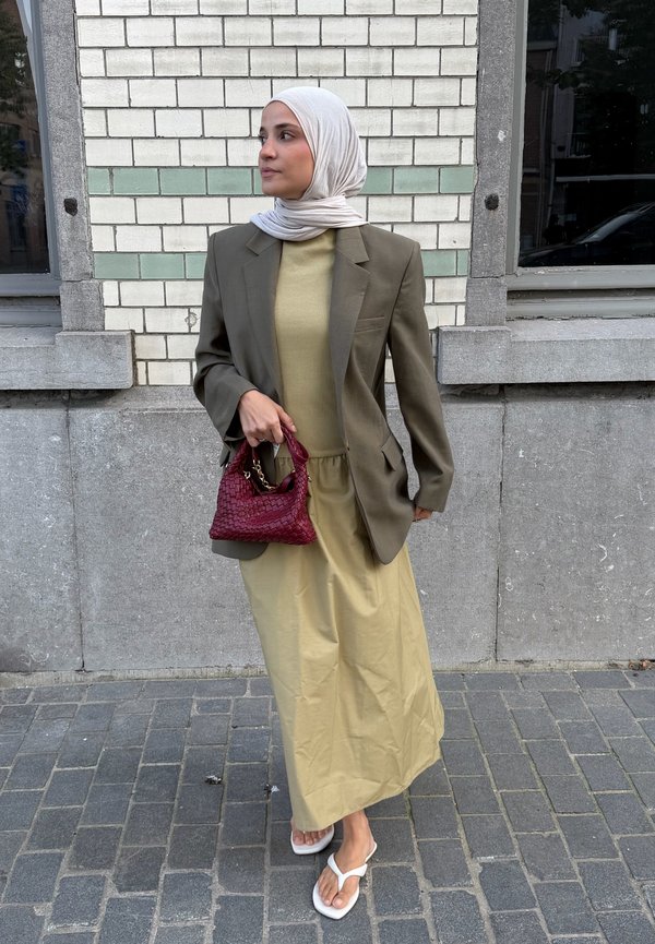 Femme portant un hijab blanc, un blazer olive, une robe beige, des sandales blanches, tenant un sac à main rouge, debout dans une rue pavée près d'un mur en briques.