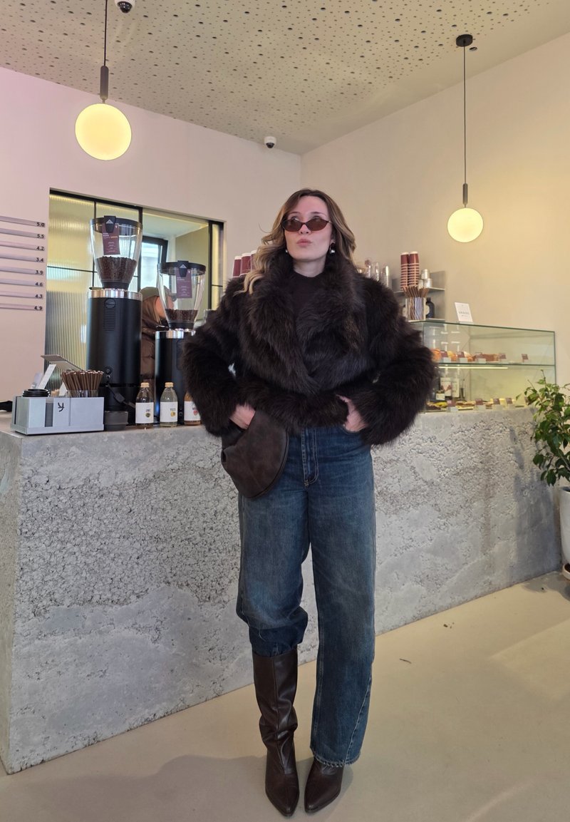 Cappotto di pelliccia marrone, maglia marrone scuro, jeans larghi blu e stivali marroni fino al ginocchio. Accessori includono occhiali da sole tondi e colorati e una borsa marrone.