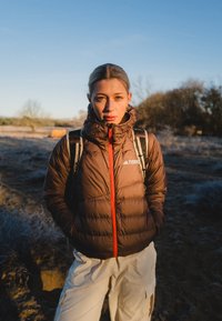 Jeune femme debout en plein air dans un champ givré, portant une doudoune marron et un pantalon beige avec un sac à dos sous un ciel bleu clair.