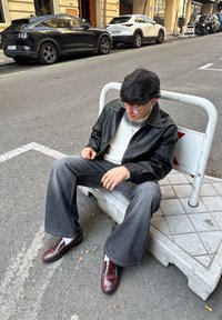 Veste en cuir noire sur un pull crème, jeans gris évasés et chaussures bordeaux. Assis sur une barrière en béton dans un cadre urbain.