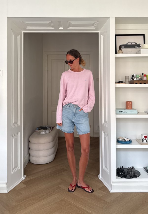 Vrouw met een zonnebril, een roze trui, een spijkershort en rode slippers staat in een witte kamer met planken en een houten vloer.