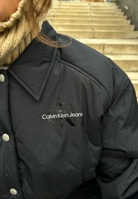 Fekete puffkabát fényes "Calvin Klein Jeans" logóval és "CK" felirattal a mellkason, látható gombokkal és textúrázott gallérral.
