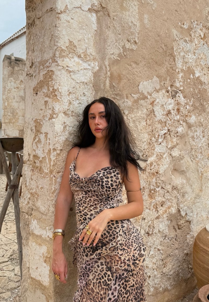Vestito con stampa leopardata, scollo a cowl, corpetto aderente e orlo con volants sovrapposti. Indossato con accessori dorati accanto a un muro di pietra rustico.