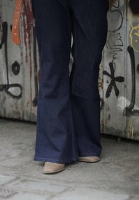 Calças de ganga de brim azul escuro com corte flare, combinadas com sapatos de bico fino em tom bege claro; cinto de couro castanho visível com múltiplos buracos.