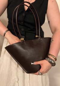 Bolsa tote de couro castanho com duas alças longas, forma estruturada e ferragens em tom de ouro. Textura suave com detalhes de costura mínimos.