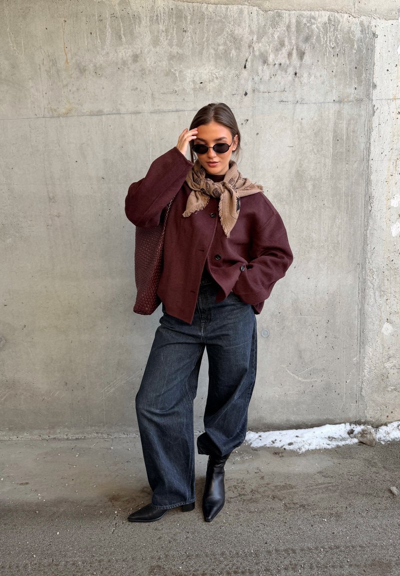 Femme portant une veste oversize bordeaux, une écharpe beige, un jean noir à jambes larges, des bottines noires et des lunettes de soleil, debout contre un mur en béton.