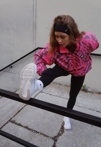 Ryškiai rožinė ir pilka raštuota striukė, juodos tamprės, balti sportbačiai. Asmuo tempia koją per juodą turėklą ant tekstūruotos dangos.