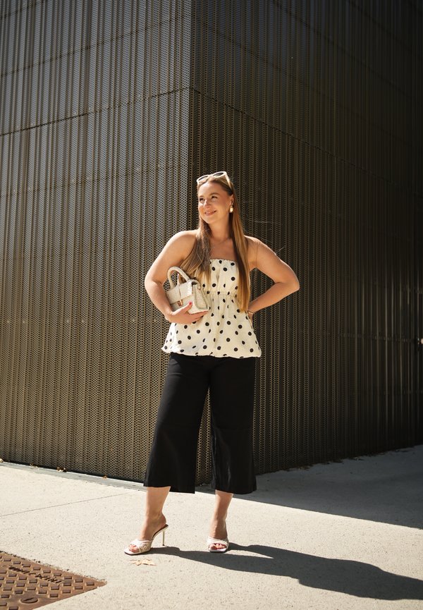 Giovane donna con occhiali da sole, top a pois, pantaloni neri e tacchi, in piedi con una piccola borsa davanti a un muro metallico testurizzato.