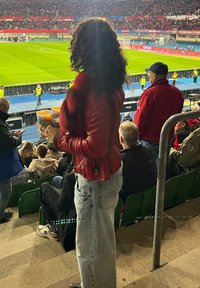 Osoba u crvenoj kožnoj jakni i plavim trapericama drži dva šarena kupa u stadionu, s nogometnim terenom i gomilom u pozadini.