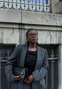 Blazer à rayures grises sur un haut noir, tenant une pochette en cuir noir. Présente un mur texturé et des détails en fer forgé en arrière-plan.