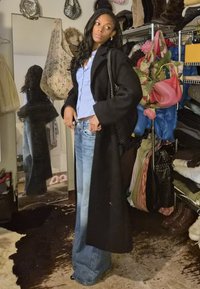 Un mannequin porte un long manteau noir par-dessus une chemise rayée bleu clair et un jean à jambes larges, avec un sac à main noir texturé, dans un placard encombré.