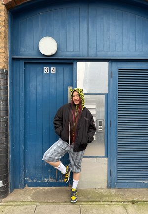 Chaqueta bomber negra sobre una sudadera gráfica con un estampado de leopardo amarillo, pantalones cortos de cuadros grises y zapatillas amarillas y azules. El fondo presenta una puerta azul.