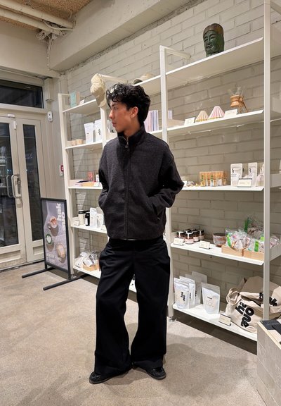 Joven con chaqueta negra y pantalones anchos negros de pie con las manos en los bolsillos dentro de una tienda con estantes blancos que sostienen productos.