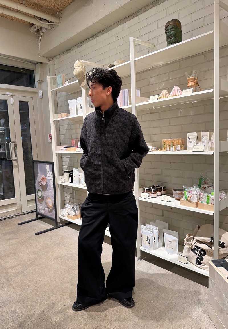 Junger Mann in schwarzer Jacke und weiten schwarzen Hosen steht mit Händen in den Taschen in einem Geschäft mit weißen Regalen, die Produkte halten.