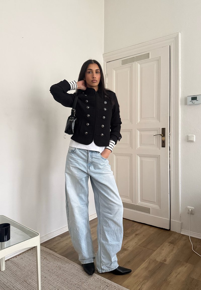 Une femme se tient à l'intérieur, portant une veste noire à double boutonnage, un chemisier à manches longues rayé, un jean à jambes larges clair, des bottes noires et un sac à main noir.