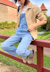 Καναβάς καφέ μπλούζα πάνω από ένα λευκό μπλουζάκι, μπλε τζιν γενναιόδωρα και καφέ σουέτ παπούτσια χωρίς κορδόνια, καθισμένος σε ξύλινη κολώνα σε εξωτερικό χώρο.