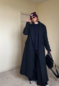 Personne portant un long manteau noir, un pull sombre, un pantalon large noir, des baskets argentées, un bonnet rayé, des lunettes de soleil noires, tenant un sac noir à l'intérieur.