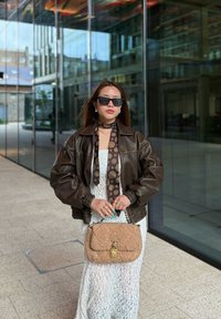 Bruine leren bomberjack, witte kanten jurk, gebloemde sjaal, zwarte zonnebril en een beige nepbont handtas met een gouden sluiting.